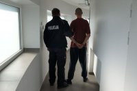 policjant trzyma za rękę zatrzymanego. Zatrzymany ma założone kajdanki. Stoją obok celi.