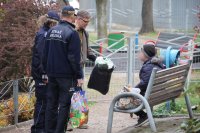 policjantka pracownik ops i strażnik wręczają paczkę bezdomnej na ławce w parku