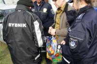 bezdomny w kurtce z napisem &quot;Rybnik&quot; odbiera paczkę z rąk pracownika ops. Obok policjantka i strażnik miejski
