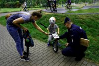 policjant zapina odblask na rowerek dziecka. na rowerku siedzi dziecko w kasku a obok stoi kobieta